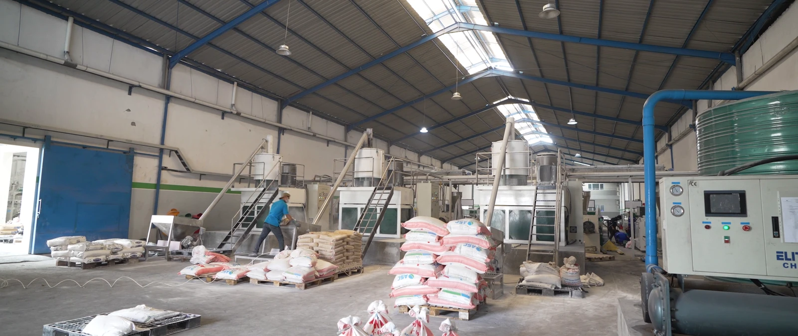 Area produksi pabrik dengan mesin, saluran pipa, dan bahan baku.